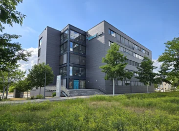 Außenansicht des neuen Standorts am Würzburger Hubland: dunkles Bürogebäude mit verglastem Treppenhaus und türkisfarbenem Eingang